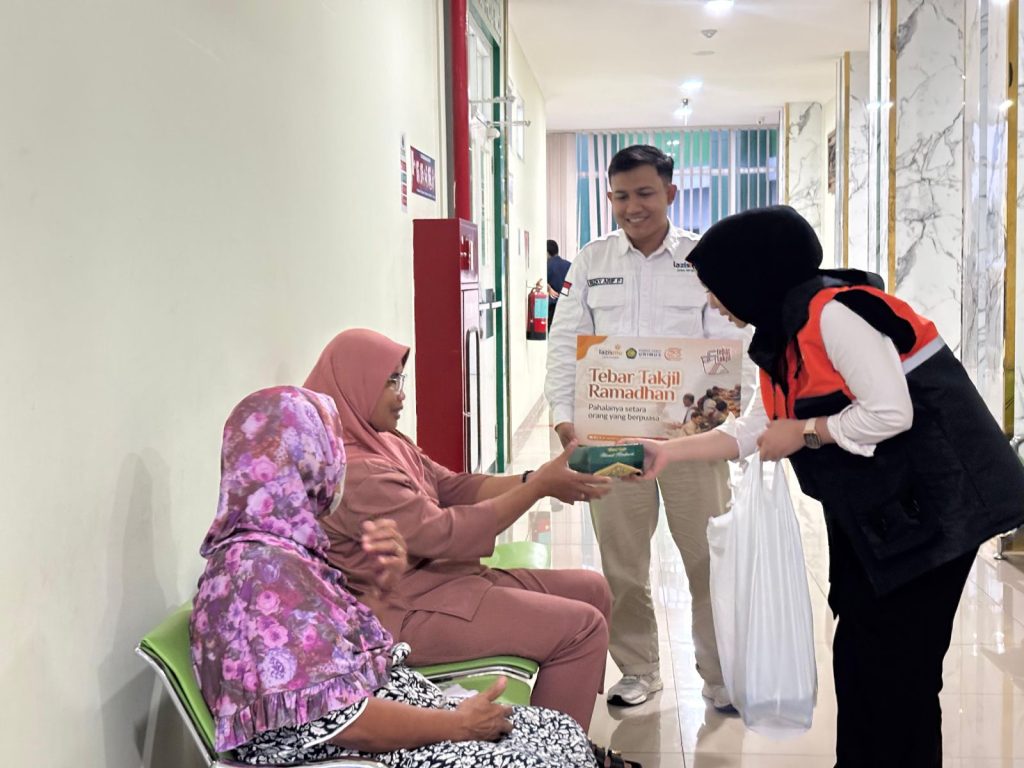 Seorang relawan wanita Lazismu berrompi oranye memberikan kotak takjil kepada seorang wanita paruh baya berjilbab merah muda di koridor rumah sakit yang bersih.